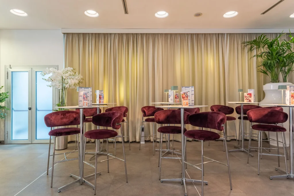 Nell'immagine il dettaglio di tavolini e sedie. Su ogni tavolo delle lampade e il menù del bar. L'area è circondata da delle tende e arredata da piante.