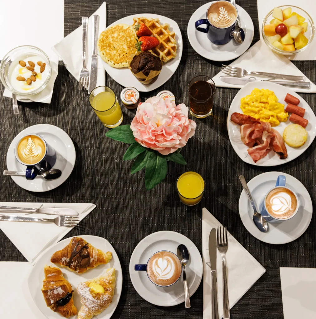 Nell' immagine un tavolo allestito per la prima colazione. Piatti salati come bacon e uova strapazzate. Proposte dolci: brioche, pancake, muffin, macedonia e yogurt. Completano il tavolo spremute d'arancia e cappuccini.
