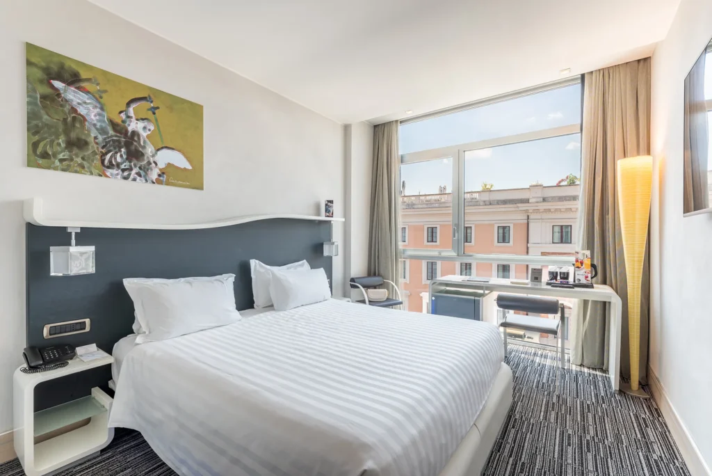 Pictured is the double bed and desk with a view of the city of Rome. On the wall opposite the bed a television set.