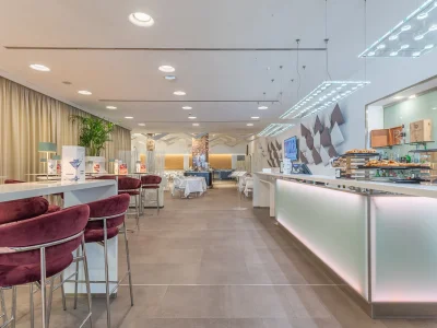 The picture shows the Hotel Bar area. The picture shows the detail of the tables and chairs. On each table are lamps and the bar menu. The area is surrounded by curtains and furnished with plants. You can also see the counter with a small buffet.