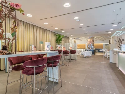 The picture shows the Hotel Bar area. The picture shows the detail of the tables and chairs. On each table are lamps and the bar menu. The area is surrounded by curtains and furnished with plants.