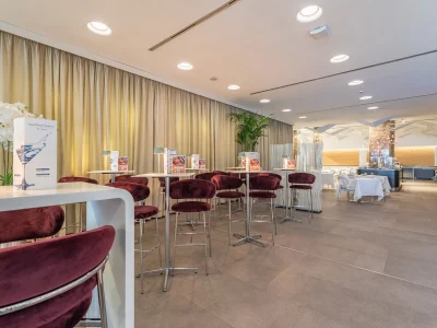 The picture shows part of the bar counter and the detail of the tables and chairs. On each table are lamps and the bar menu. The area is surrounded by curtains and furnished with plants.