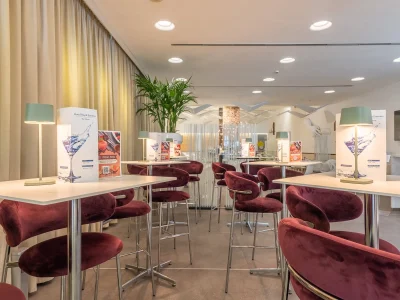 The picture shows the detail of tables and chairs. On each table are lamps and the bar menu. The area is surrounded by curtains and furnished with plants.