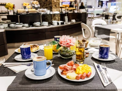 Pictured is a table set up for breakfast. Savoury dishes such as bacon and scrambled eggs. Sweet options: brioche, pancakes, muffins, fruit salad and yoghurt. Orange juices and cappuccinos complete the table. The buffet table can be seen in the background.