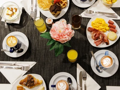 Pictured is a table set up for breakfast. Savoury dishes such as bacon and scrambled eggs. Sweet options: brioche, pancakes, muffins, fruit salad and yoghurt. Orange juices and cappuccinos complete the table.