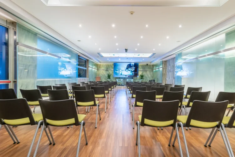 Nell'immagine la Sala Lucrezia del Centro Congressi dell' Hotel Royal Santina, vista dal fondo della sala. La disposizione della sala è a platea, divisa in due settori e ogni fila è composta da quattro sedie. Nella sala è presente anche un tavolo per i relatori con microfoni e dietro un ledwall.