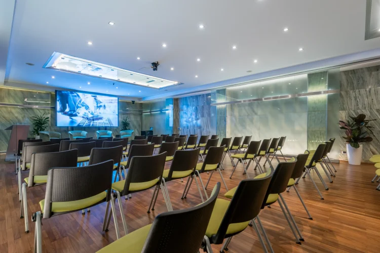 Nell'immagine la Sala Lucrezia del Centro Congressi dell' Hotel Royal Santina, vista dal fondo della sala. La disposizione della sala è a platea, divisa in due settori e ogni fila è composta da quattro sedie. Nella sala è presente anche un tavolo per i relatori con microfoni e dietro un ledwall.