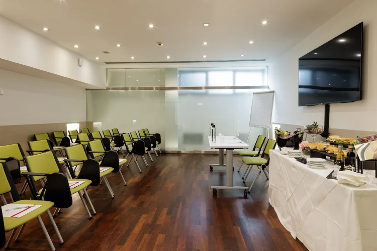 Nell'immagine la Sala Bianca del Centro Congressi dell' Hotel Royal Santina. La disposizione della sala è a platea. Davanti un tavolo per i relatori con due postazioni, uno schermo e una lavagna a fogli. Su un lato della sala è presente anche un buffet per i partecipanti.