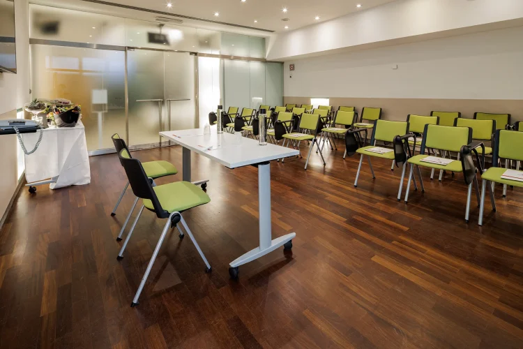 Nell'immagine la Sala Bianca del Centro Congressi dell' Hotel Royal Santina. La disposizione della sala è a platea. Davanti un tavolo per i relatori con due postazioni.