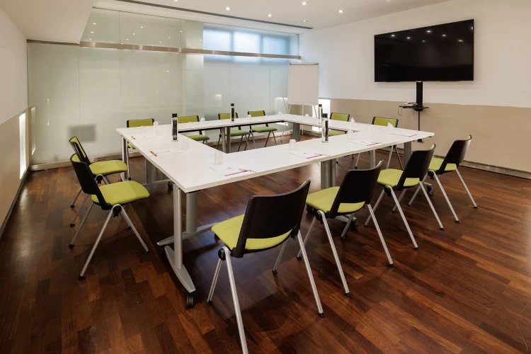 Nell'immagine la Sala Bianca del Centro Congressi dell' Hotel Royal Santina. La disposizione della sala è a tavolo unico. Il tavolo è quadrato ed è posizionato al centro della sala, circondato dalle sedie per i partecipanti. Nella sala ci sono anche: uno schermo per le proiezioni e una lavagna a fogli.