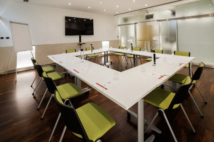 Nell'immagine la Sala Bianca del Centro Congressi dell' Hotel Royal Santina. La disposizione della sala è a tavolo unico. Il tavolo è quadrato ed è posizionato al centro della sala, circondato dalle sedie per i partecipanti. Nella sala ci sono anche: uno schermo per le proiezioni e una lavagna a fogli.