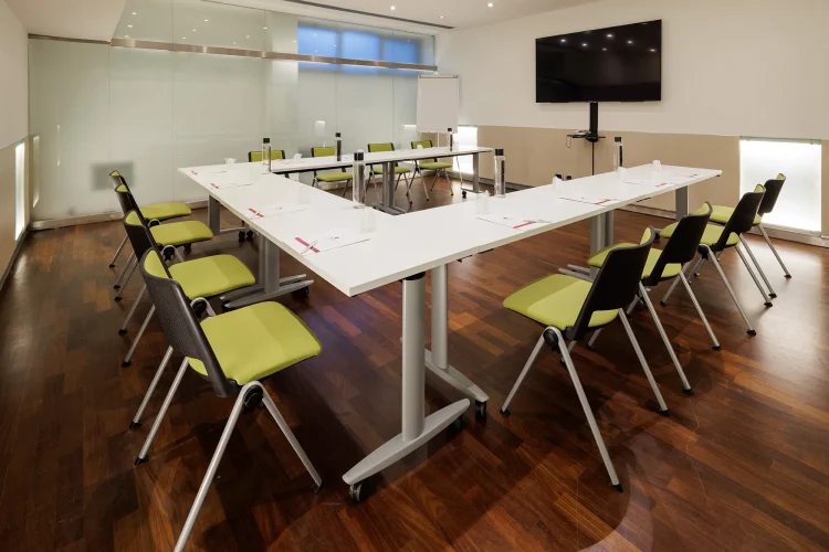 Nell'immagine la Sala Bianca del Centro Congressi dell' Hotel Royal Santina. La disposizione della sala è a ferro di cavallo: il tavolo è nel centro della sala con attorno le sedie per i partecipanti. Nella sala ci sono anche: uno schermo per le proiezioni e una lavagna a fogli.