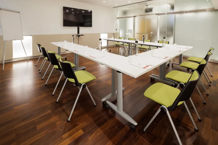 Nell'immagine la Sala Bianca del Centro Congressi dell' Hotel Royal Santina. La disposizione della sala è a ferro di cavallo: il tavolo è nel centro della sala con attorno le sedie per i partecipanti. Nella sala ci sono anche: uno schermo per le proiezioni e una lavagna a fogli.