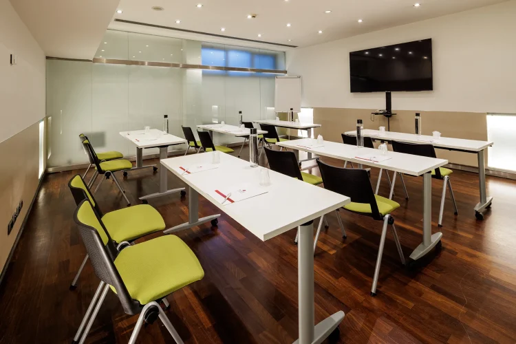 Nell'immagine la Sala Bianca del Centro Congressi dell' Hotel Royal Santina, vista in diagonale. La disposizione della sala è a banchi di scuola. Per ogni banco ci sono due sedie. Nella sala ci sono anche: uno schermo per le proiezioni e una lavagna a fogli.