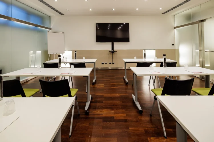 Nell'immagine la Sala Bianca del Centro Congressi dell' Hotel Royal Santina. La disposizione della sala è a banchi di scuola. Per ogni banco ci sono due sedie. Nella sala ci sono anche: uno schermo per le proiezioni e una lavagna a fogli.