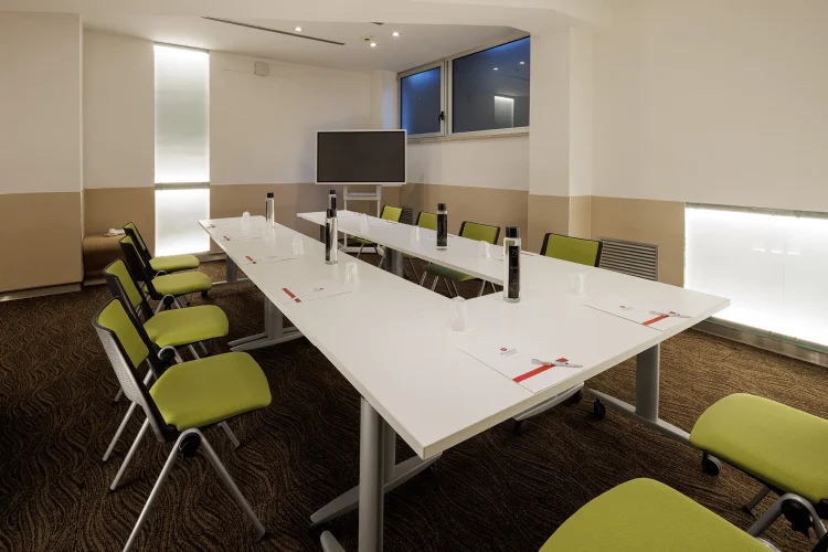 Nella foto, la sala riunioni Priscilla ha una disposizione a ferro di cavallo con dieci postazioni. Su un lato c'è anche uno schermo per le proiezioni.